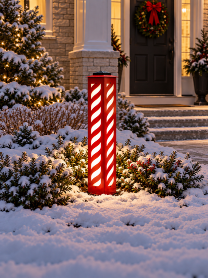 A red metal post outdoor holiday light with white diagonal stripes stands in a snow-covered garden in front of a house porch with a wreath on the door.