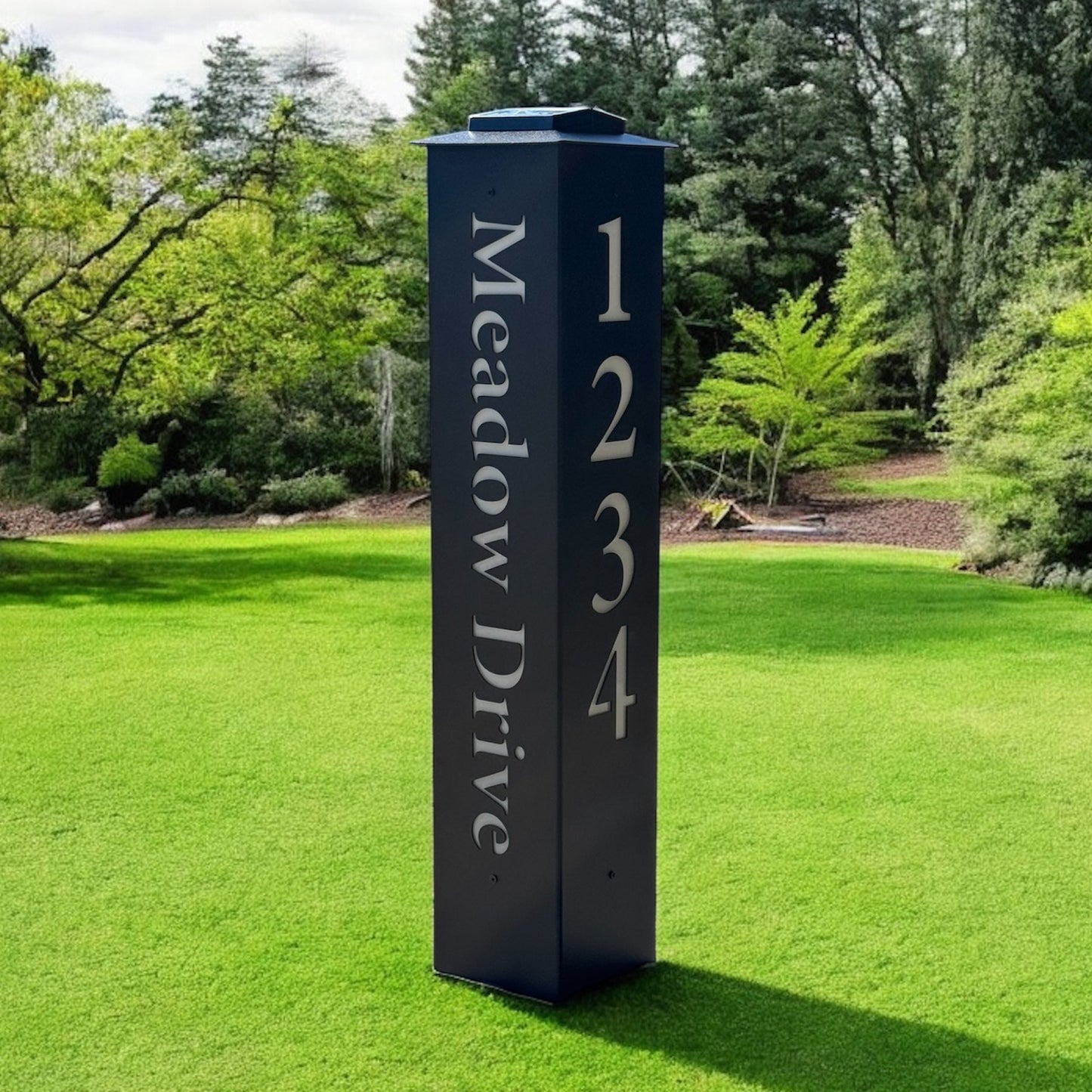 Black address signpost with 'Meadow Drive' and numbers on a grassy lawn with trees in the background