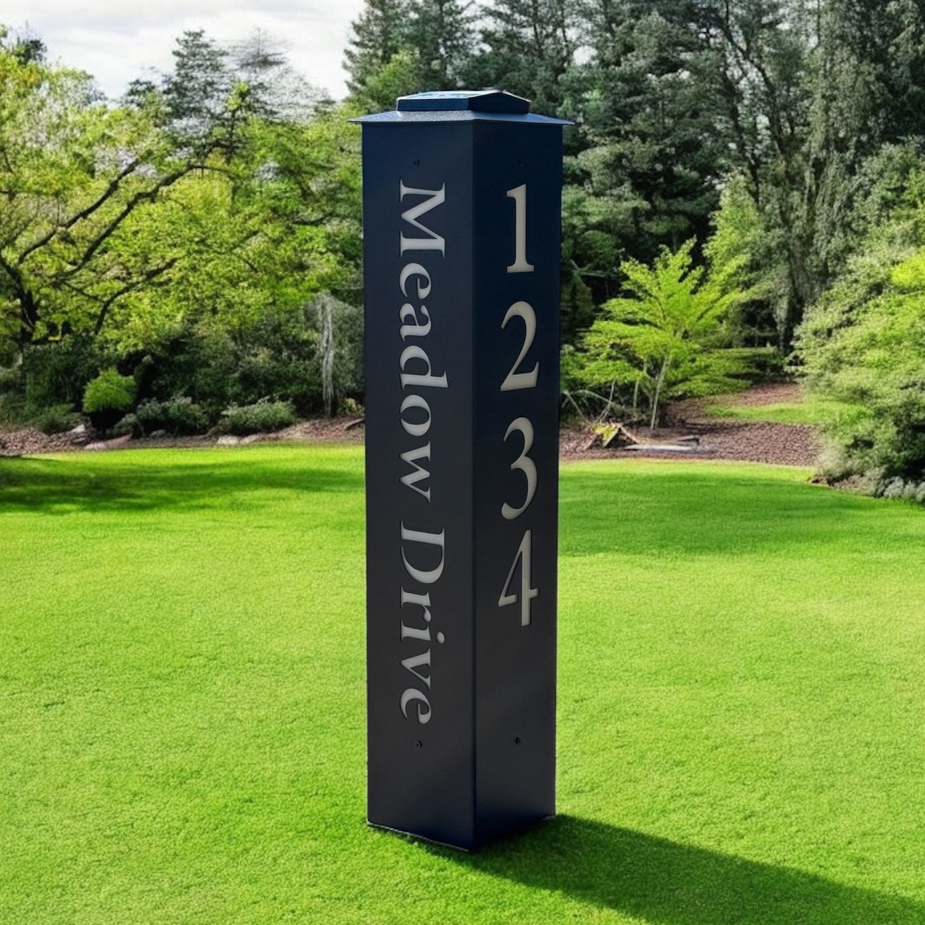Black address signpost with 'Meadow Drive' and numbers on a grassy lawn with trees in the background
