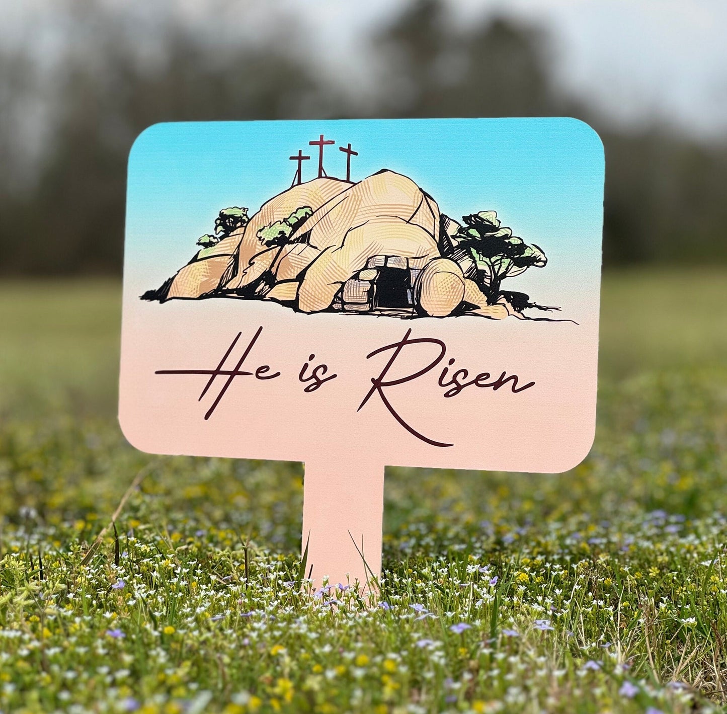 He is Risen Easter Stake with three crosses on a hill, decorative sign for Easter yard decor in a grassy field.