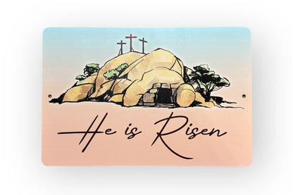 Close-up of 'He is Risen' Easter metal sign showing tomb and crosses with gradient background.