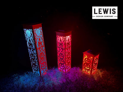 Three tall rectangular LED lanterns with intricate cutout patterns, shown lit in red, pink, and orange hues on grass at night. The brand logo 'LEWIS DESIGN COMPANY' is visible in the top right corner.