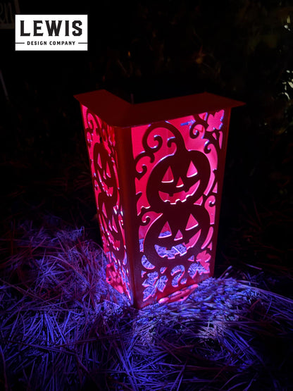 A red Halloween-themed decorative lantern with intricate pumpkin cutouts, glowing with pink and purple light in a dark outdoor setting on straw.