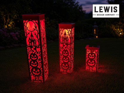 Three red LED solar-powered Halloween yard lanterns of different heights with laser-cut jack-o'-lantern and haunted house patterns, illuminated on a grassy lawn at dusk. A 'LEWIS DESIGN COMPANY' logo is visible in the top right corner.